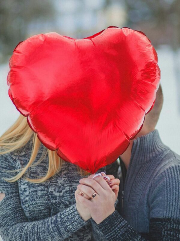 A couple embraces behind a heart-shaped balloon outdoors, depicting love and romance.